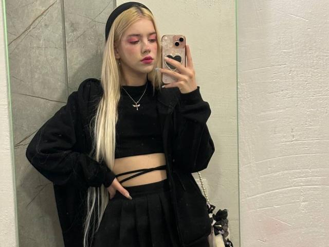 ShawtyLola with long blonde hair, black beanie, pink eyeshadow, black crop top, black jacket, black skirt, hand in pocket, mirror selfie, phone with heart case, white walls.