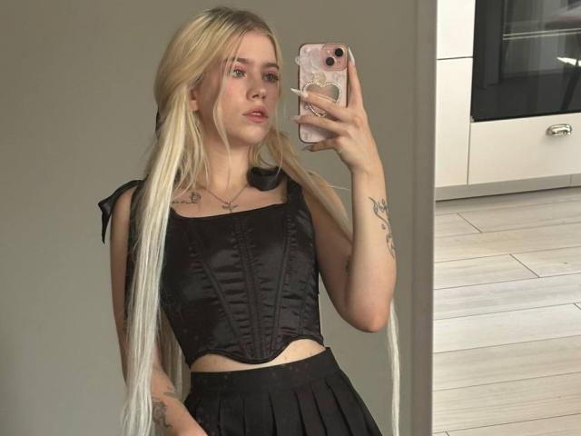 ShawtyLola with long blonde pigtails, black corset, and black skirt, holding phone in mirror selfie. Boobs visible under corset. Tattoo on left arm. Neutral expression.