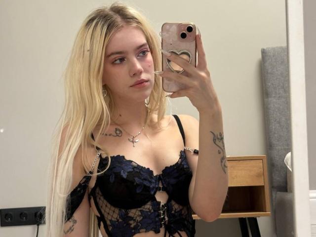 ShawtyLola with long blonde hair, wearing black lace lingerie with floral patterns, holding a pink phone, showing tattoos on her arm and chest, taking a mirror selfie.