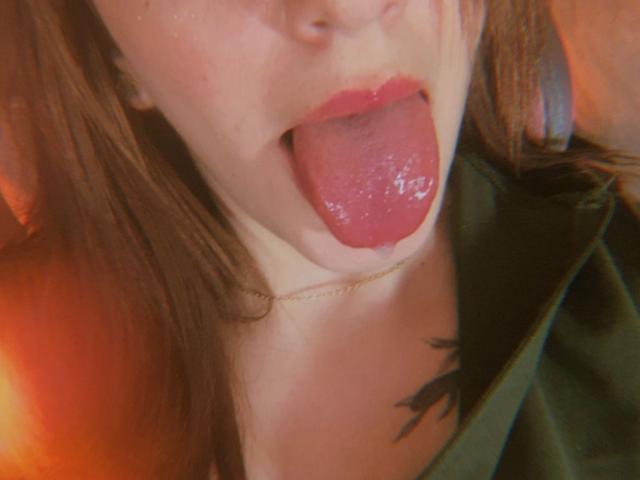 LiaHarrys with tongue out, red lips, and a small gold chain, showing a black tattoo near her neck, wearing a green jacket. Her tongue is wet and shiny.