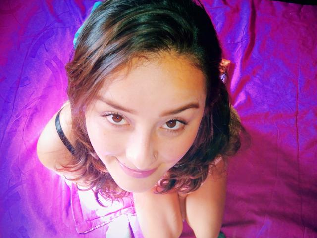 ViollettKim with wavy brown hair, brown eyes, and a nose ring, smiling up at the camera, wearing a black top, kneeling on a purple cloth.