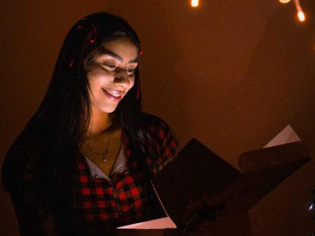 JessicaSoler with long dark hair, red checkered shirt, holding a book, smiling, light brown skin, small red cross necklace, dimly lit with warm string lights.