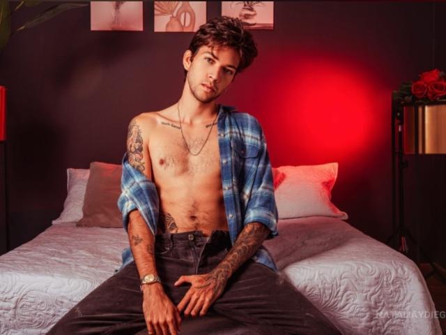 Shirtless HottersTeam with tattoos, blue plaid shirt, black pants, sitting on bed, red light behind, wristwatch, serious expression, dark curly hair, slight beard, chest tattoo, right hand on crotch.