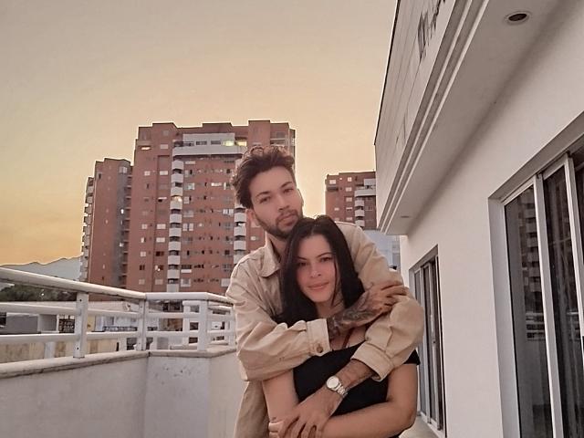 HottersTeam with brown hair and beard, wearing beige shirt, hugging HottersTeam with black hair and tattoos, wearing black top, on rooftop at sunset. Both have cute, relaxed expressions.