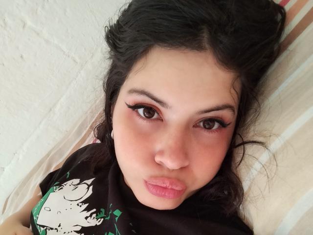 HottersTeam has dark wavy hair, brown eyes with long lashes, pink lipstick, and is wearing a black shirt with a green and white graphic. Lying down, pouty expression.