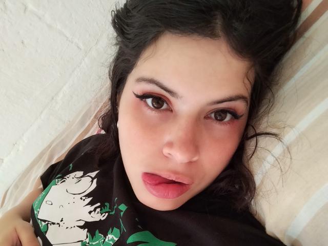 HottersTeam with dark wavy hair, pink lipstick, and thick black eyeliner, lying down, wearing a black shirt with green and white graphic, looking directly at the camera.