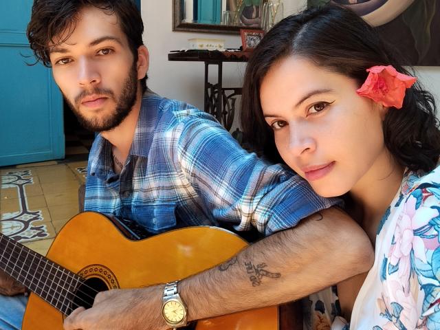 HottersTeam with brown hair, beard, and blue plaid shirt holds acoustic guitar, tattoo on forearm. HottersTeam with pink flower in hair, tan skin, wearing floral shirt, leans on him.