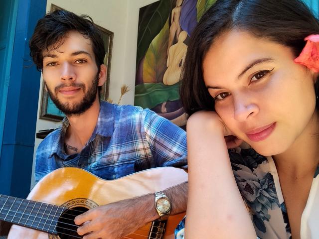 HottersTeam, a cute brunette with a pink flower in her hair, rests her head on her arm, smiling. HottersTeam, a bearded guy with a guitar, wears a blue shirt.