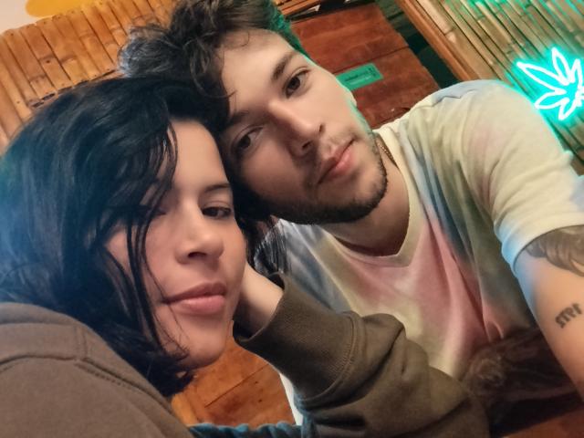 HottersTeam with black hair and pink lips leans on HottersTeam with curly brown hair and beard, wearing a tie-dye shirt, in a wooden room with neon light.