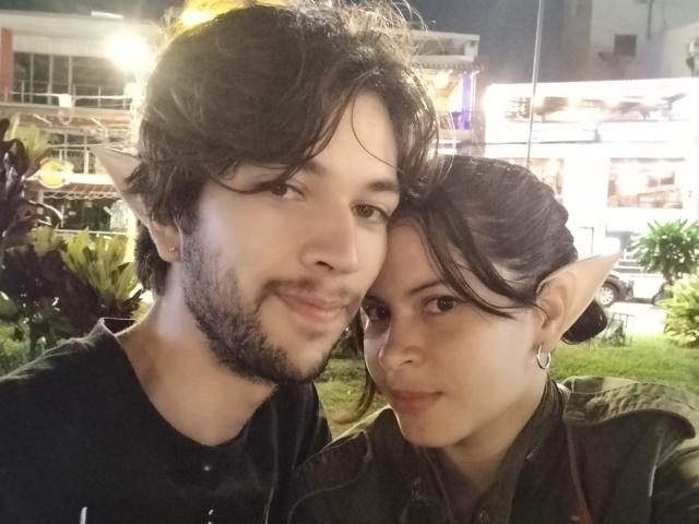 HottersTeam with elf ears, both have pointy ears, dark hair, and light skin, smiling at the camera, guy with beard, girl with earrings, night setting, casual clothes.
