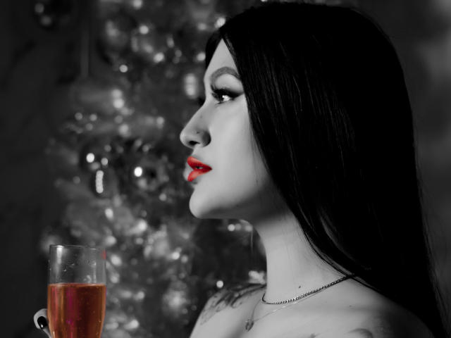 EmmaRosseti with black hair, red lipstick, holding a glass of red wine, white Christmas tree in the blurred background, visible tattoo on right shoulder, black-and-white photo.