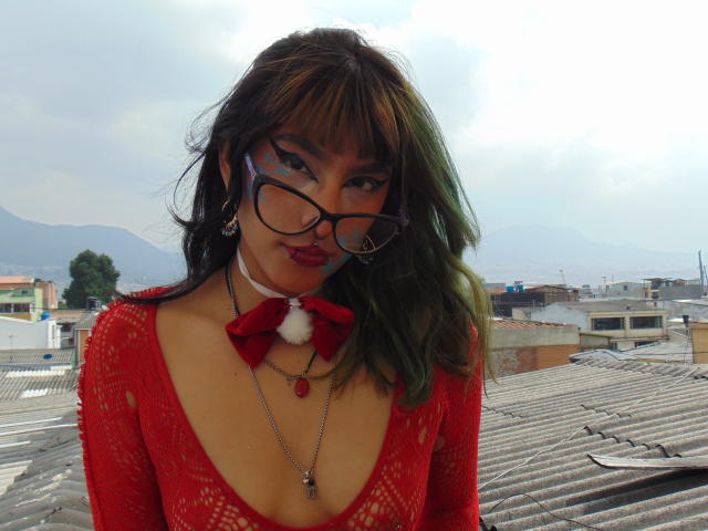 SerineHotAss con cabello verde y marrón, top de encaje rojo, escote pronunciado, gafas negras, lazo rojo, pompon blanco, piercing en la nariz y collar verde, de pie en la azotea..
