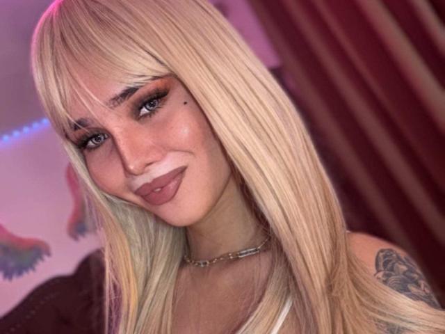 LoveMeKara with blonde hair, blue eyes, and a tattoo on her shoulder, wearing a choker, smiling with full lips. Her tits are small, and she's cute.