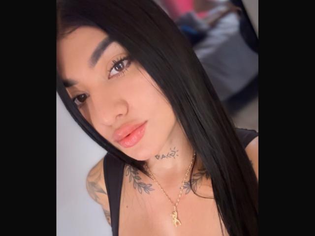 KittySwt with long black hair, full pink lips, tattoo under chin, tattoos on chest, wearing black top, gold necklace, brown eyes, slightly parted lips, looking at camera.