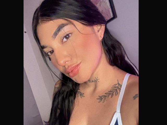 KittySwt with black hair, brown eyes, and pinkish blush, wearing a white top, showing tattoos on neck and chest, pouty lips, and subtle smile.