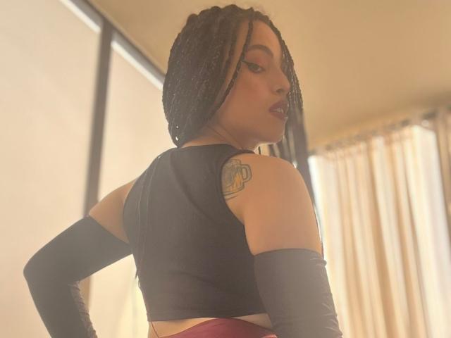 ElizaLovely with braided hair, black crop top, black arm sleeves, red panties, and a tattoo on her right shoulder, looking over her shoulder with a sultry expression.