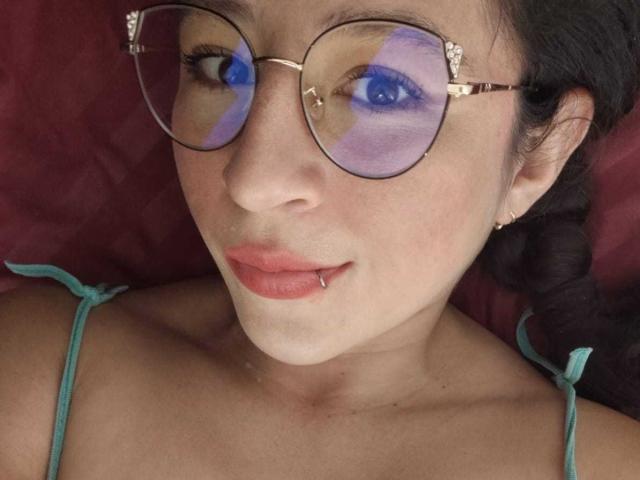 SweetyHot69 with glasses reflecting blue and purple eye makeup, has a lip piercing and a braid, wearing a green-strapped top, with fair skin and red lips.