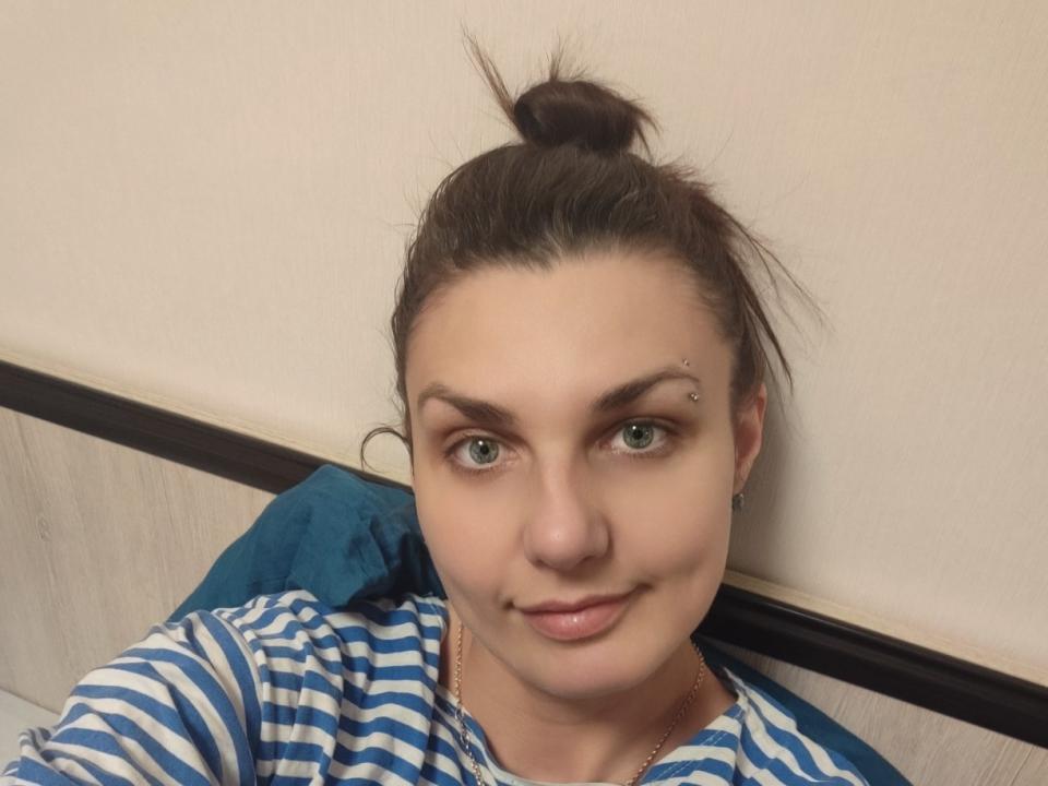 MagicSmile69 with a topknot, green eyes, and a small facial piercing on the right eyebrow, wearing a blue and white striped shirt, smiling subtly.