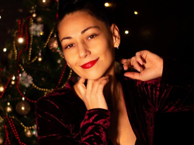 LiluGrants with red lipstick, smiling, wearing a velvet purple dress, her hands near her neck, dark hair in a bun, Christmas lights in the background, warm lighting.