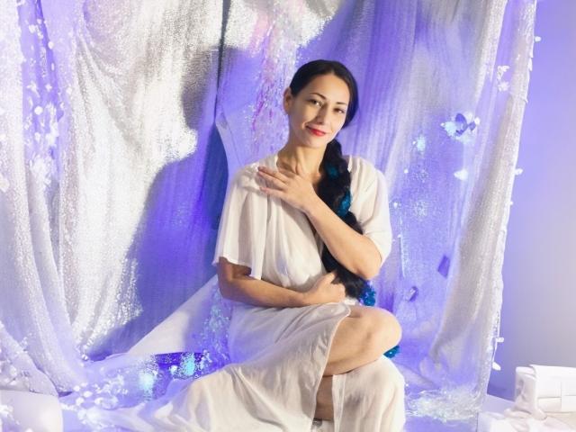 LiluGrants, with dark hair in a braid, sits in a white dress, covering her small tits with one hand, smiling, against a purple-lit, glittery backdrop.