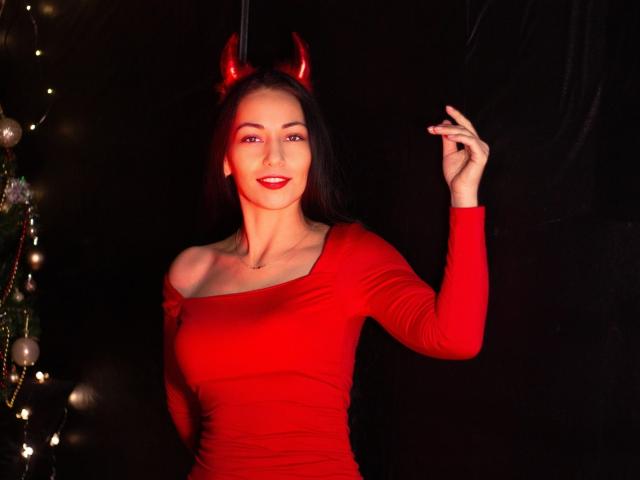 LiluGrants with red devil horns, wearing a tight red off-shoulder top, smiling, holding up a finger, red lipstick, dark hair, medium tits, dark background, Christmas tree on left.