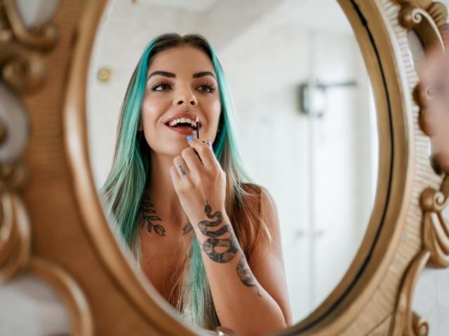 BruCute with teal hair, tattoos on arms, and a cute smile, applying blue lipstick in an ornate gold mirror. Her tits are not visible. Her ass is not visible.