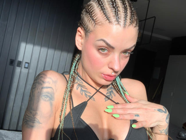 BruCute with braided hair, green nails, tattoos, pink blush, and a black top, showing off her tits and a hand near her chest. Bright light on her face.