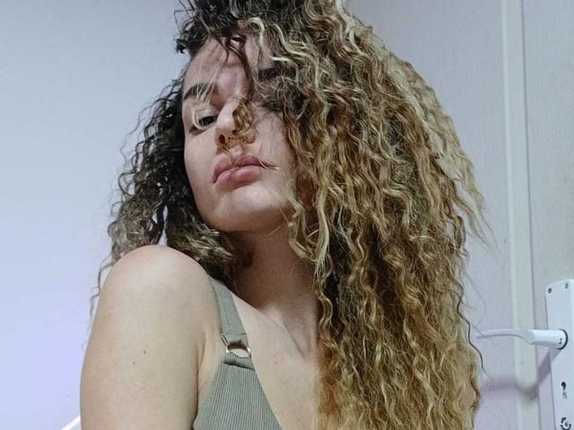 IngridI has curly blonde and brown hair, wearing a green tank top, with a nose piercing and a slight pout, looking down at the camera.