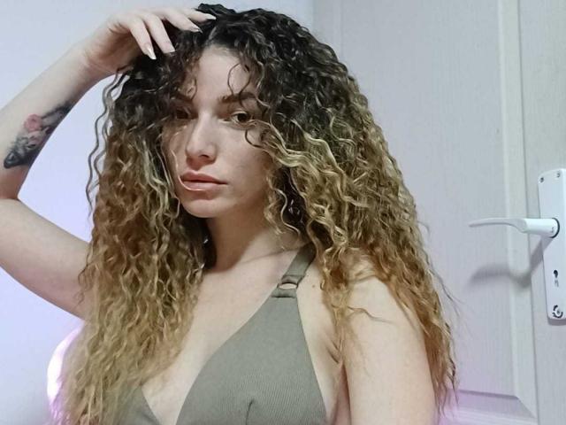 IngridI with curly brown and blonde hair, wearing a grey halter top, has her hand in her hair, showing a tattoo on her left arm, and a slight pout.