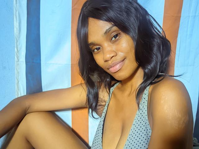 Tiah with dark wavy hair, cute nose ring, and subtle smile, wearing a grey polka dot top, sitting with knee up, showcasing her small, perky tits.