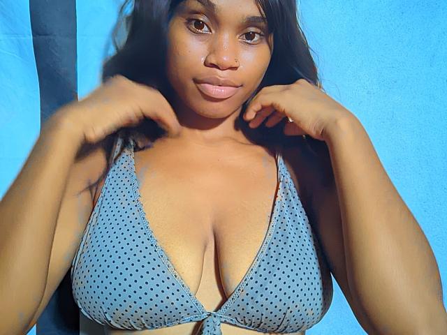 Tiah has dark brown skin, long black hair, and is wearing a blue polka dot bikini top, showing her big, round boobs. She's holding her hair with both hands.