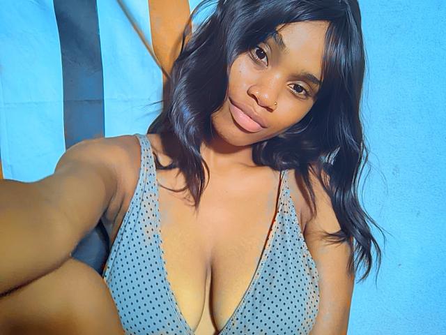 Tiah with medium brown skin, wavy black hair, and small nose, wearing a grey polka-dot bikini top, showing her large tits and smiling softly at the camera.