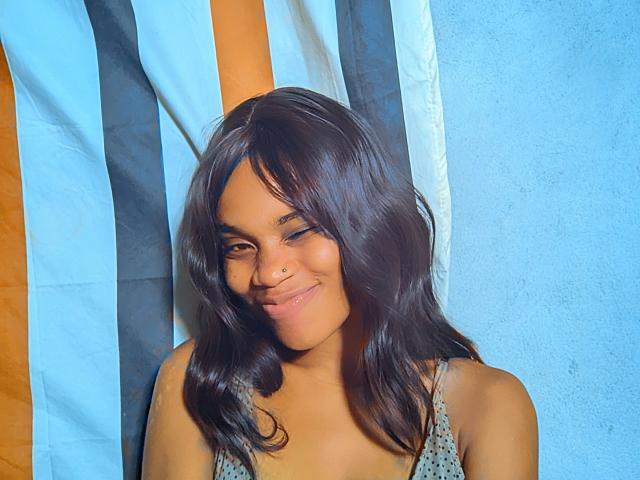 Tiah has medium brown skin, wavy dark brown hair, and a cute nose ring. She's smiling with a slight blush, wearing a green polka dot top.