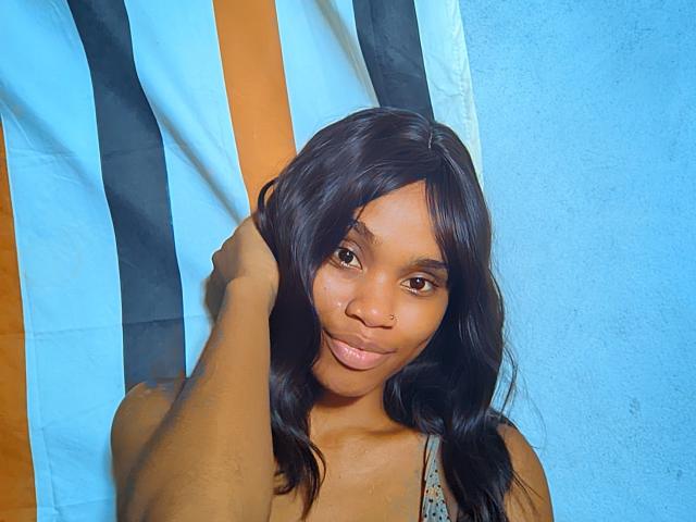 Tiah with dark wavy hair, brown eyes, and nose ring, holding her hair, wearing a patterned top, smiling slightly, standing in front of a blue and black striped flag.