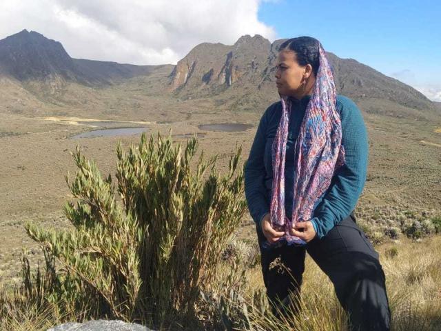 BantuRousse stands in a rugged landscape, wearing a teal long-sleeve shirt, black pants, and a colorful scarf, with hands on her hips, looking right. Mountain and lake in background.