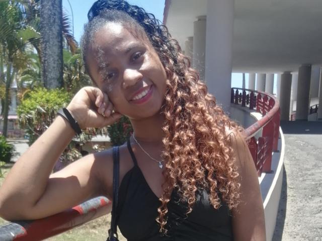 Edrwina69 with long curly brown hair, black tank top, smiling, sunlight on face, wearing a necklace, black bracelet, hand on cheek, red and white railing, outdoor setting.