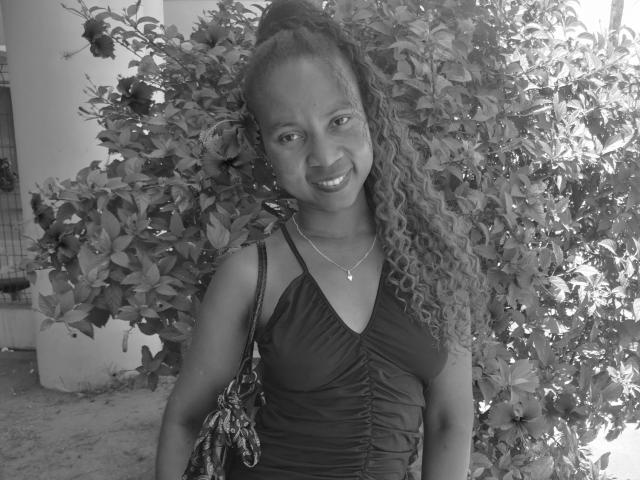 Edrwina69 with curly hair, smiling, wearing a ruched black dress, necklace, and patterned bag, standing in front of a leafy bush, black-and-white photo.