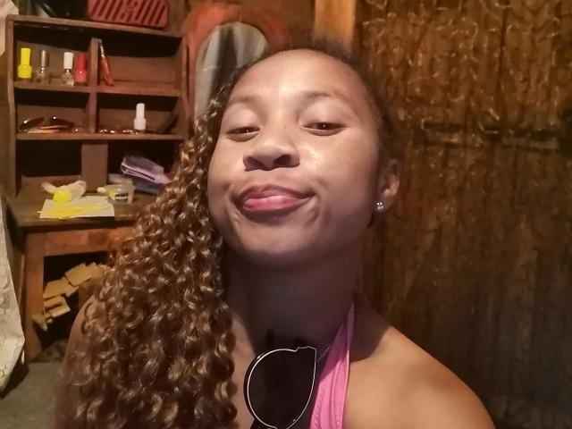 Edrwina69 with dark brown skin, curly brown hair, and pink halter top, sticking out pink tongue, wearing small diamond earrings, in dimly lit room with wooden textures.