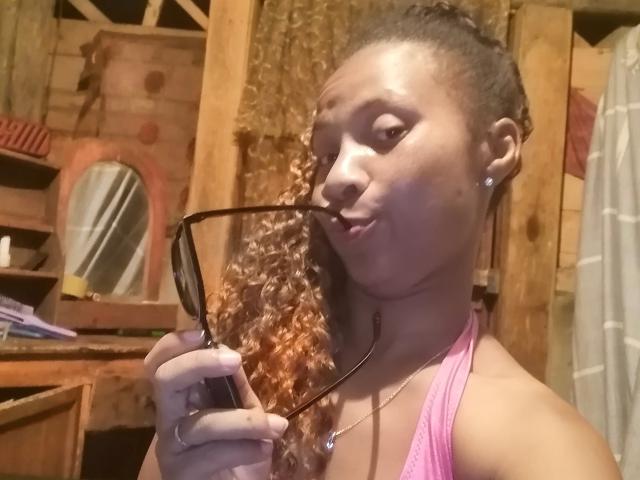 Edrwina69 with curly brown hair, pink tank top, holding black glasses, pouting, wearing diamond earrings, in a wooden room. Her tits are visible.
