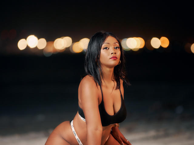 MayaRichh has dark skin, black shoulder-length hair, and is wearing a black sports bra with white Calvin Klein underwear, showing her large tits and curvy ass. Night photo.