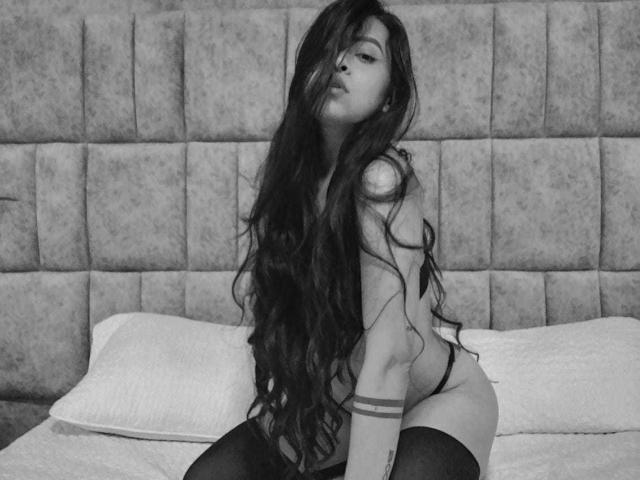 Paulinna Rodriguez, met lang, golvend donker haar, draagt zwarte knighthoogtes en een zwart stringbroekje, zittend op een bed, waarbij haar billen en kleine borsten zichtbaar zijn, met een zelfverzekerde, sexy uitdrukking..