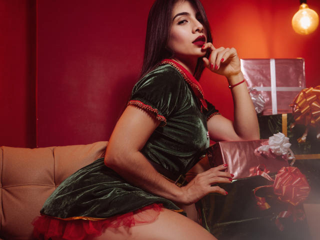 MaryLoft in a green velvet dress with red trim, biting her finger, showing her red skirt and red nails, holding a gift with a bow, on a couch.