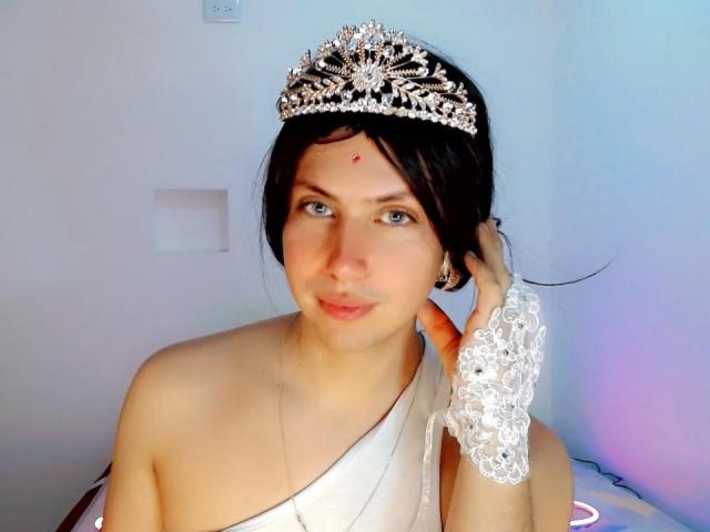 AdriudI with a jeweled tiara, white lace glove, and bare shoulders, looking at the camera, has blue eyes and a red bindi on her forehead. She's wearing a white sash.
