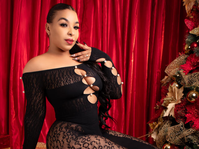 EileenBenzon with long black hair, wearing a black lace dress with cut-out circles revealing her tits, sitting next to a decorated Christmas tree, red curtains in background.