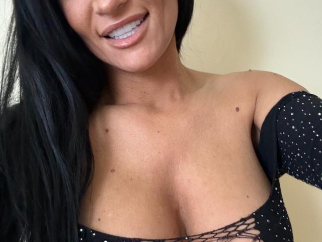 Le visage d'AndraD, souriant, laissant apparaître ses dents, ses cheveux noirs et sa peau bronzée. Elle porte un haut noir, à encolure dégagée et à lacets, qui révèle ses petites taches de beauté foncées sur sa poitrine..