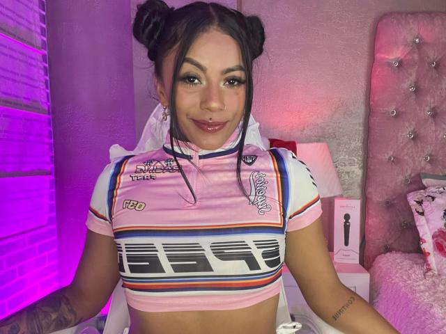 Amberv with pink and white NASCAR-style top, showing her small tits, dark hair in double buns, pink lip gloss, and tattoo on right arm, sitting in pink-lit room.