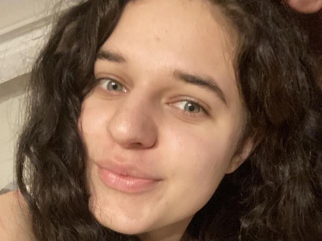 LightShannon's face, showing curly black hair, green eyes, and slightly pouty pink lips, with a light skin tone and subtle freckles, in a casual, slightly messy selfie.