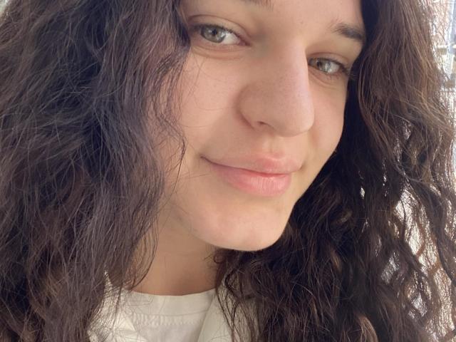 LightShannon's face with wavy brown hair, light skin, green eyes, subtle smile, and pink lips. She's wearing a white shirt, and the photo is a tight shot.