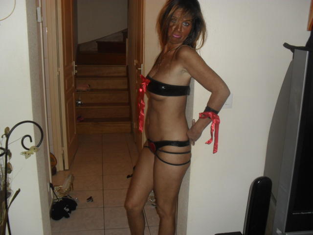 Faniae with tan skin, brown hair, and red lipstick, wearing a black latex bra and strappy black and red panties, standing in a doorway, hands on hips, looking at camera.