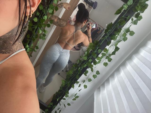 LalaVega mit Tattoos, von hinten zu sehen, trägt ein trägerloses Top und graue, eng anliegende Leggings, macht ein Selfie vor einem Spiegel und präsentiert dabei ihren Hintern, in einem Raum mit grünen Pflanzen..